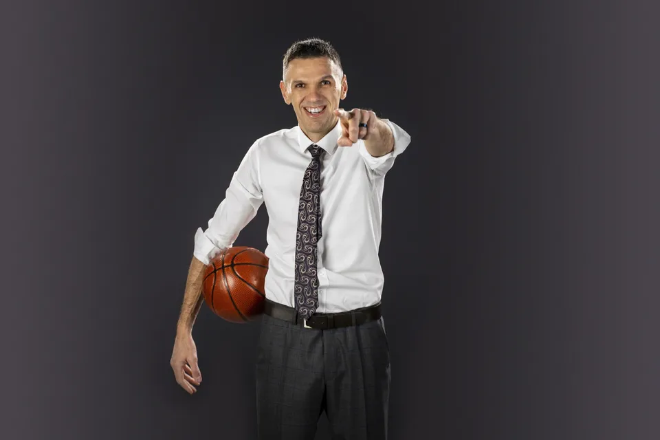 Lubo Bali in a white shirt and tie, a basketball under his arm, smiling and pointing at the camera